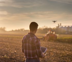 Attractive farmer navigating drone above farmland with silos and tractor in background. High technology innovations for increasing productivity in agriculture